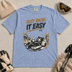 Washed denim blue t-shirt featuring a black bear resting in a hammock with ski gear and the words “Busy Taking It Easy.”