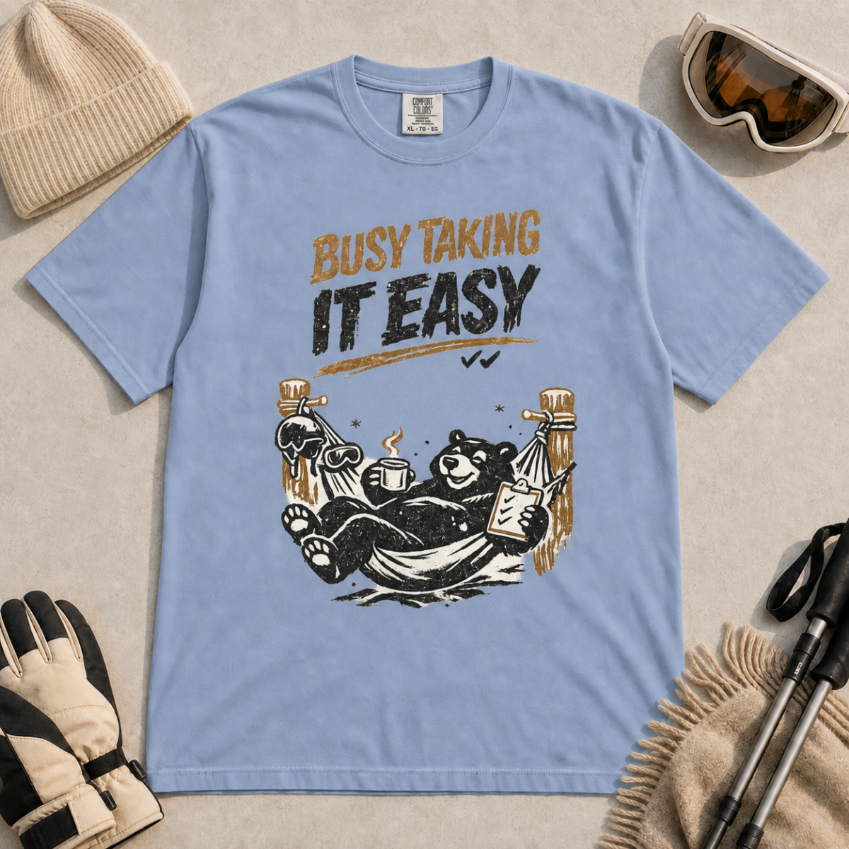 Washed denim blue t-shirt featuring a black bear resting in a hammock with ski gear and the words “Busy Taking It Easy.”