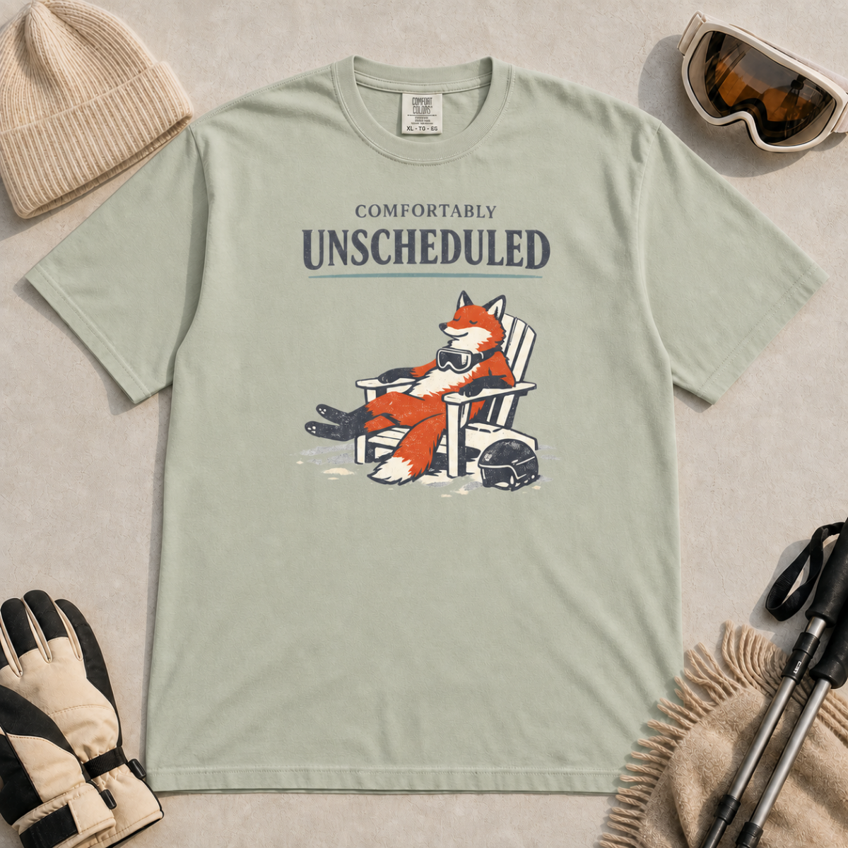 Bay green t-shirt featuring a fox relaxing in a chair beside a ski helmet with the words “Comfortably Unscheduled.”