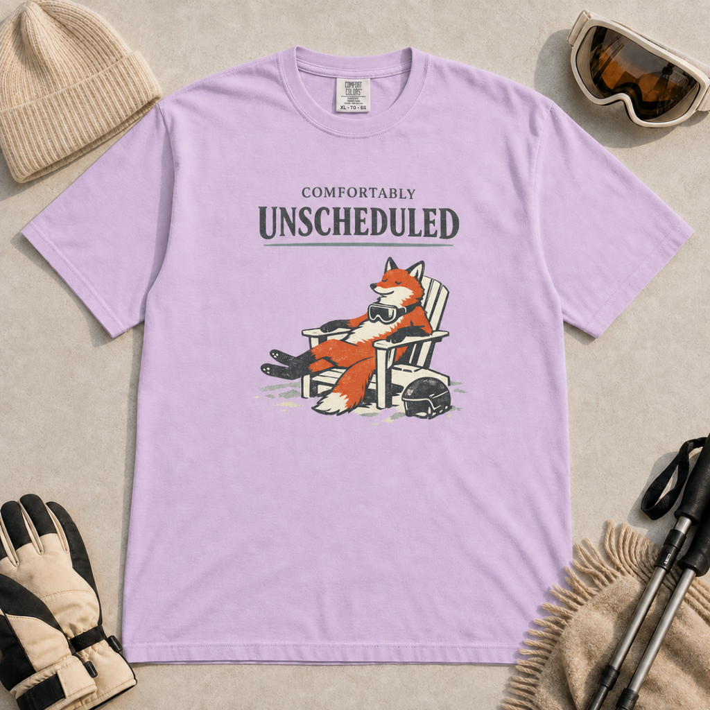 Orchid purple t-shirt featuring a fox relaxing in a chair beside a ski helmet with the words “Comfortably Unscheduled.”