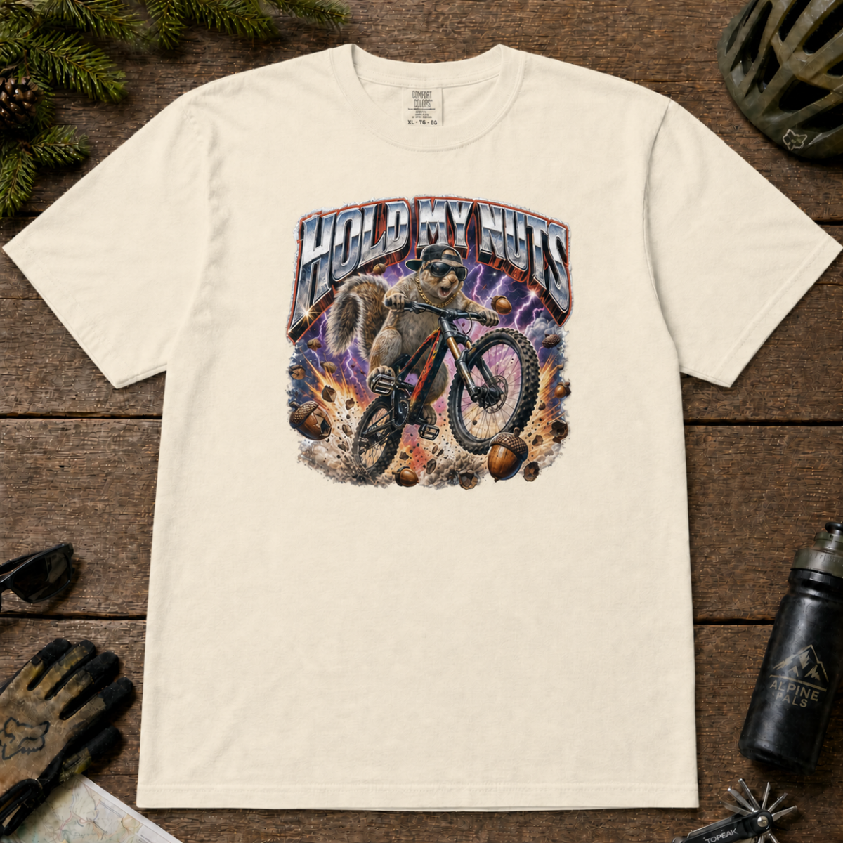 Ivory white t-shirt featuring a squirrel mountain biking through flying acorns beneath the words “Hold My Nuts.”