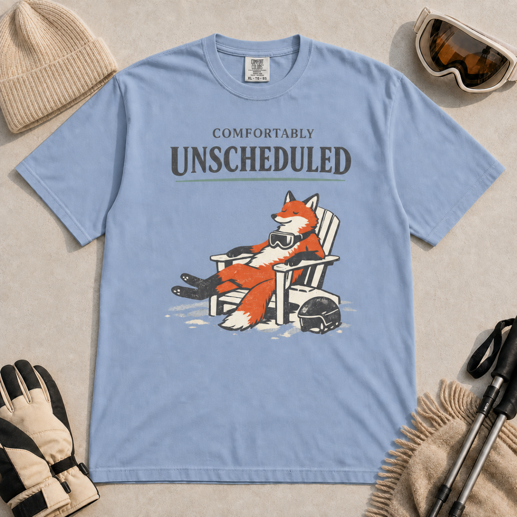 Washed denim blue t-shirt featuring a fox relaxing in a chair beside a ski helmet with the words “Comfortably Unscheduled.”