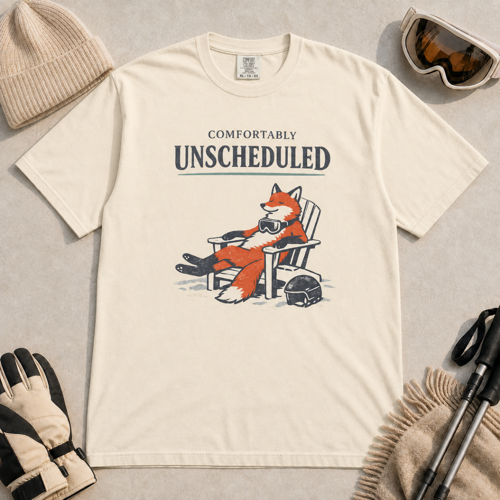 Ivory white t-shirt featuring a fox relaxing in a chair beside a ski helmet with the words “Comfortably Unscheduled.”