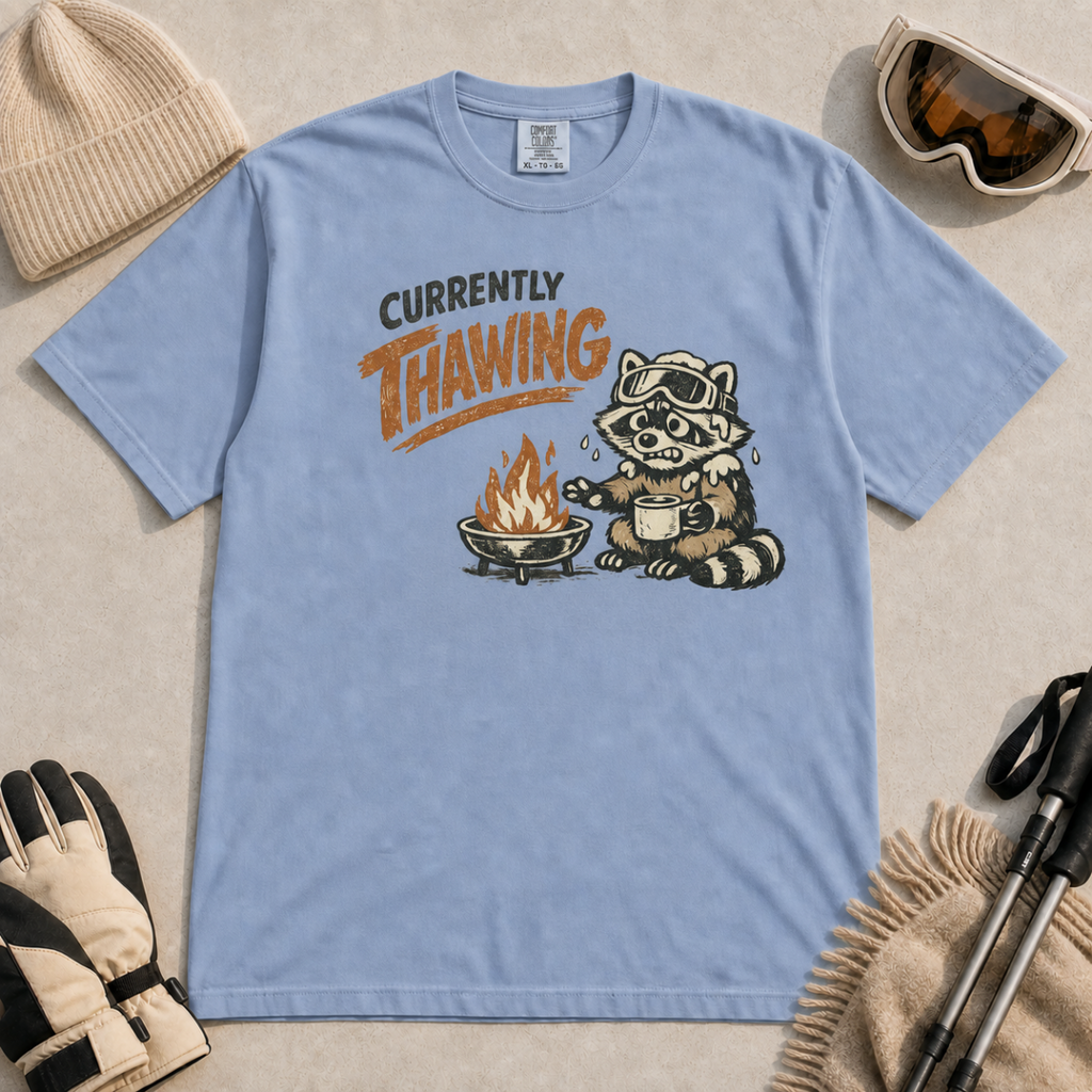 Washed denim blue t-shirt featuring a raccoon warming beside a fire with a mug and the words “Currently Thawing.”