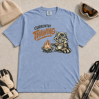 Washed denim blue t-shirt featuring a raccoon warming beside a fire with a mug and the words “Currently Thawing.”