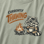 Close-up of a raccoon in ski goggles holding a mug beside a fire with the words “Currently Thawing.”