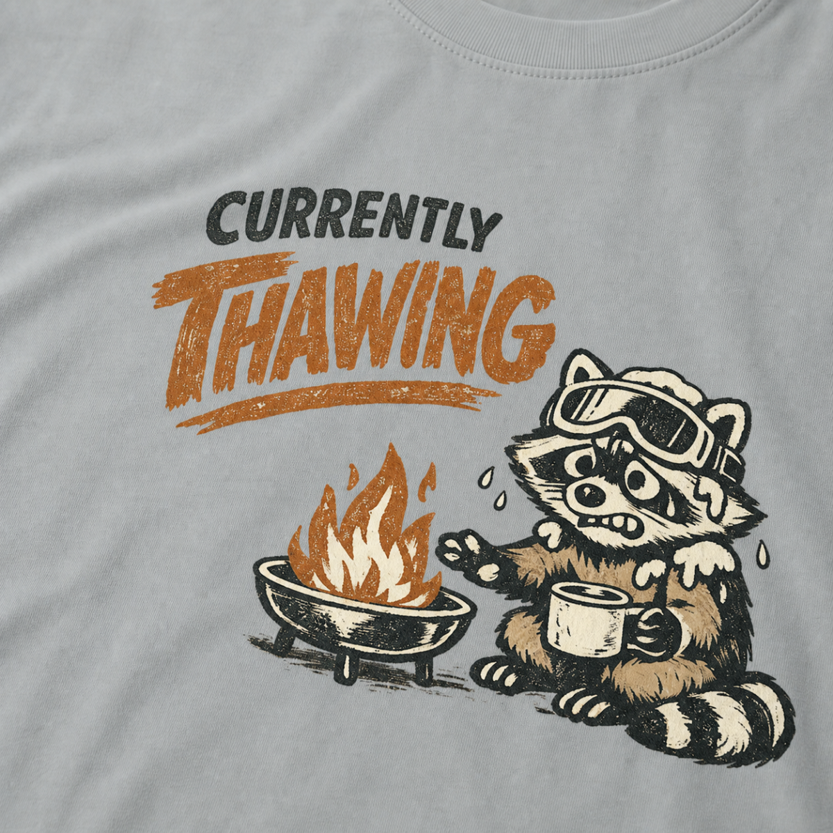 Close-up of a raccoon in ski goggles holding a mug beside a fire with the words “Currently Thawing.”