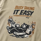 Close-up of a black bear relaxing in a ski-lodge hammock with a hot drink and checklist beneath the words “Busy Taking It Easy.”