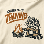 Close-up of a raccoon in ski goggles holding a mug beside a fire with the words “Currently Thawing.”