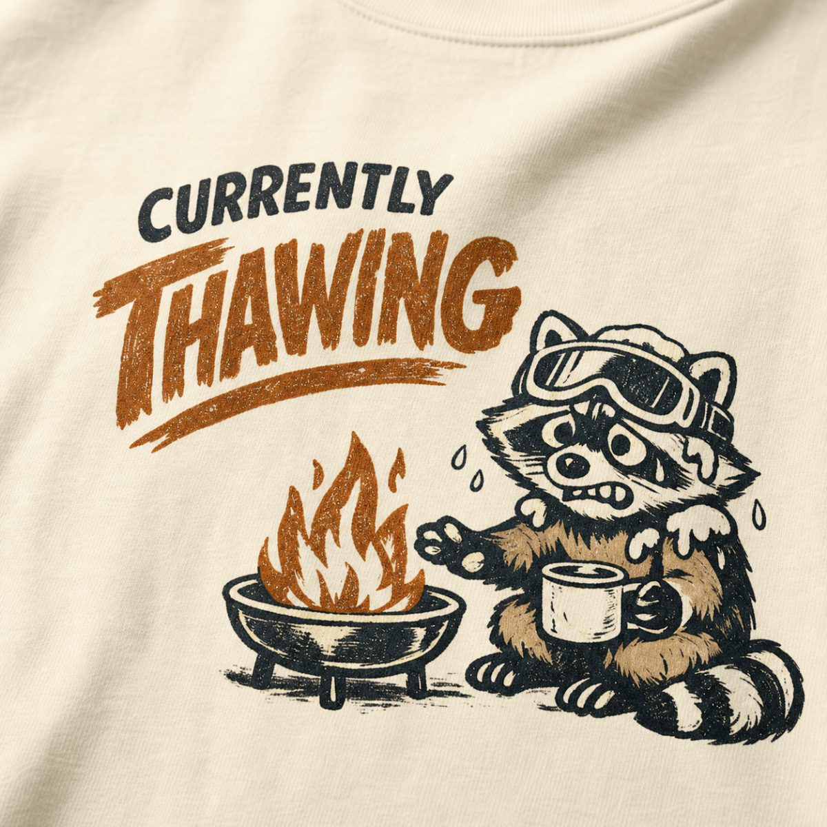 Close-up of a raccoon in ski goggles holding a mug beside a fire with the words “Currently Thawing.”