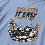 Close-up of a black bear relaxing in a ski-lodge hammock with a hot drink and checklist beneath the words “Busy Taking It Easy.”