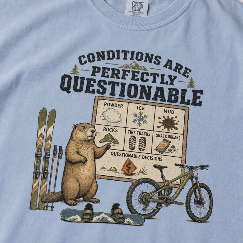 Close-up of a washed denim blue t-shirt featuring a marmot beside skis, a snowboard, a mountain bike, and a humorous conditions chart.