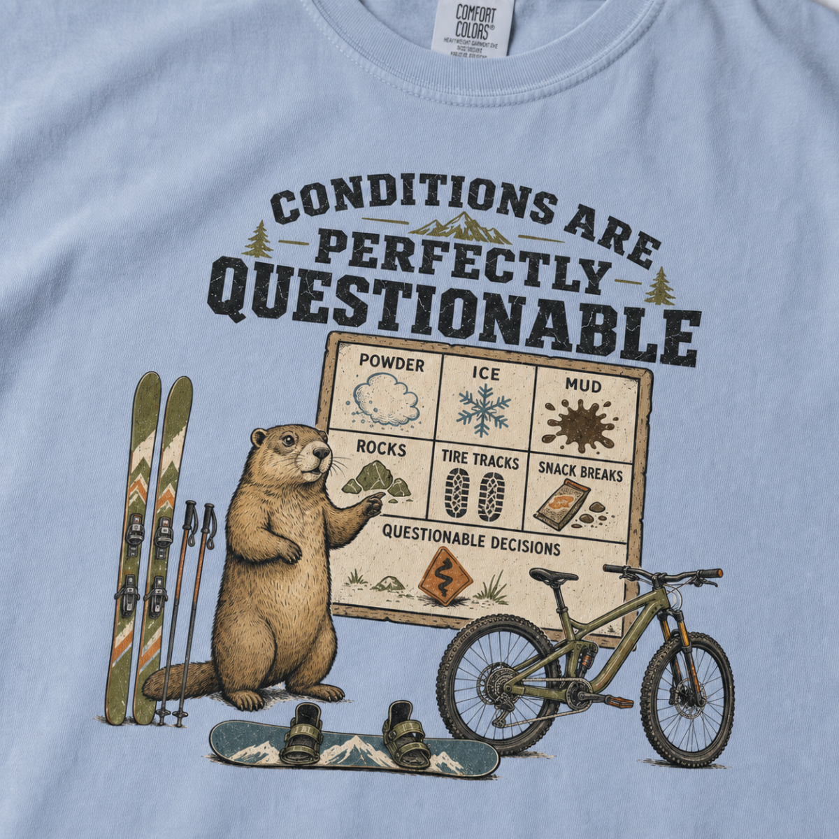 Close-up of a washed denim blue t-shirt featuring a marmot beside skis, a snowboard, a mountain bike, and a humorous conditions chart.