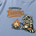Close-up of a raccoon in ski goggles holding a mug beside a fire with the words “Currently Thawing.”