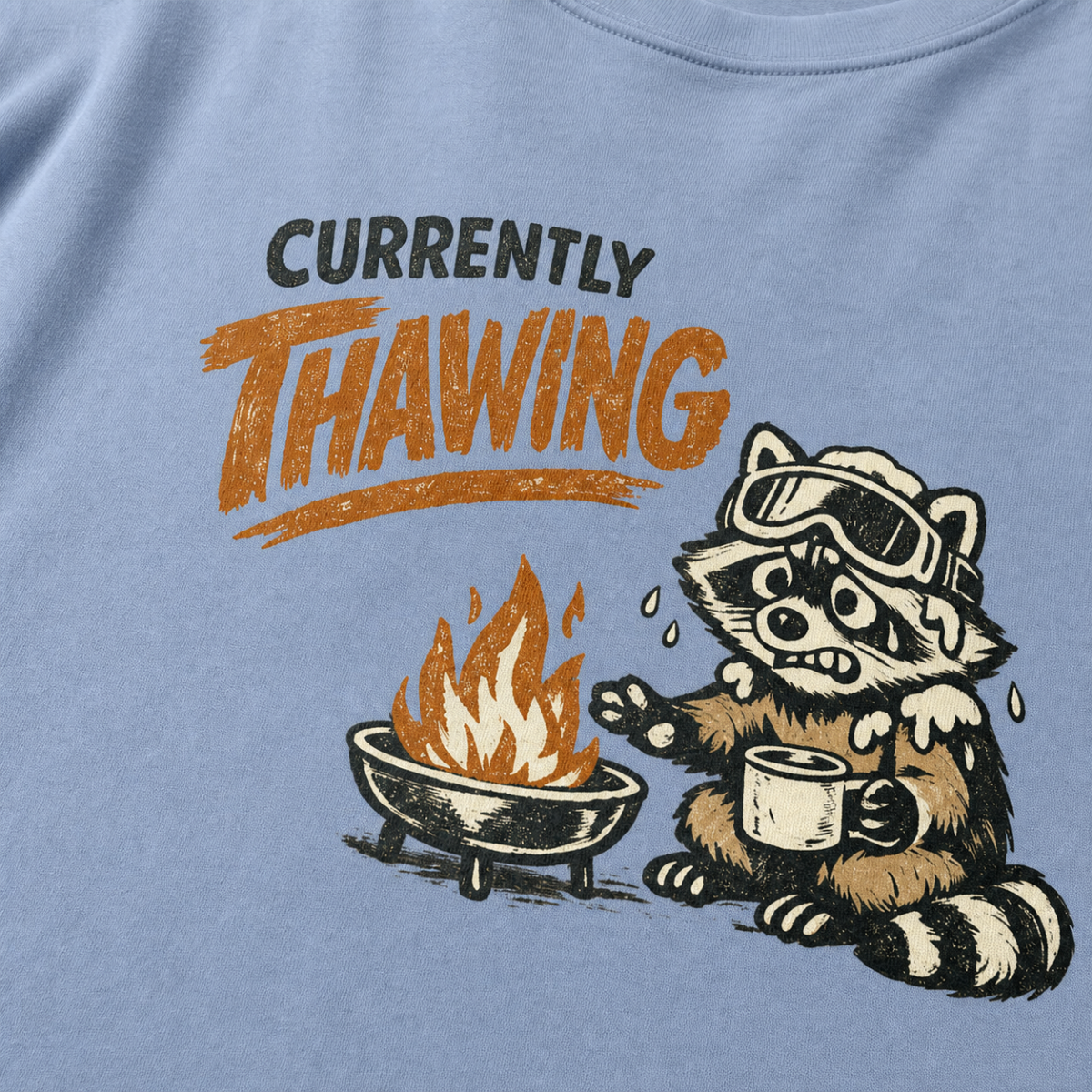 Close-up of a raccoon in ski goggles holding a mug beside a fire with the words “Currently Thawing.”