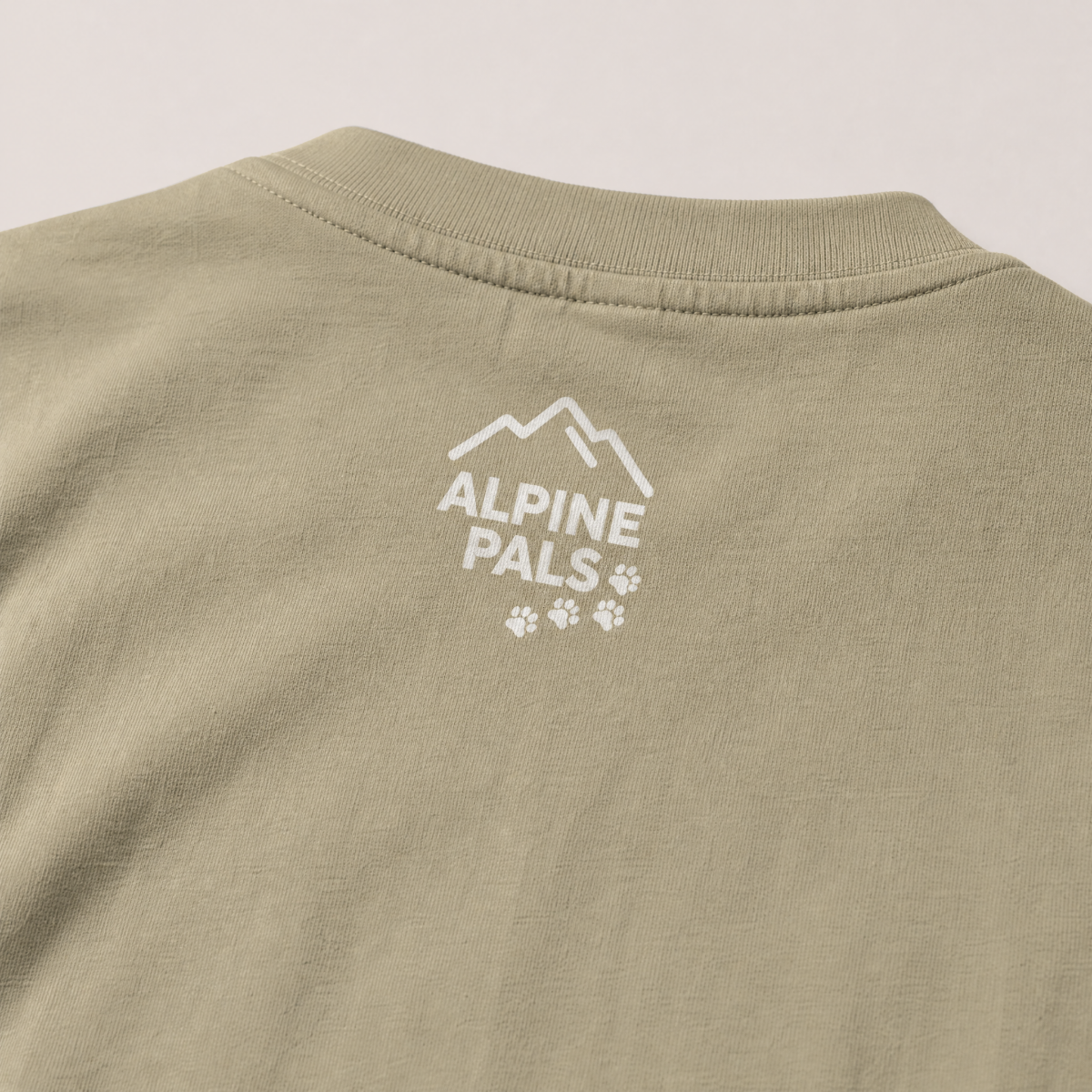 Close-up of a khaki colored shirt with 'Alpine Pals' logo on a beige background