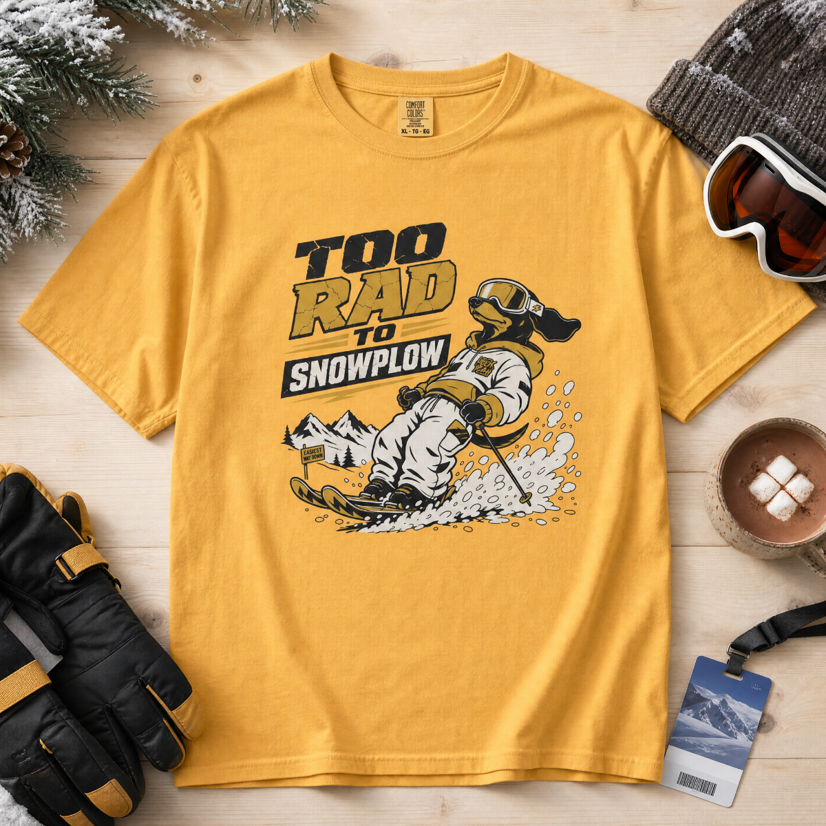 Yellow t-shirt with a graphic design of a person snowshoeing and text 'Too Rad to Snowplow' on a wooden surface with winter accessories.