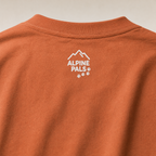 Close-up of a yam colored shirt with 'Alpine Pals' logo on a beige background
