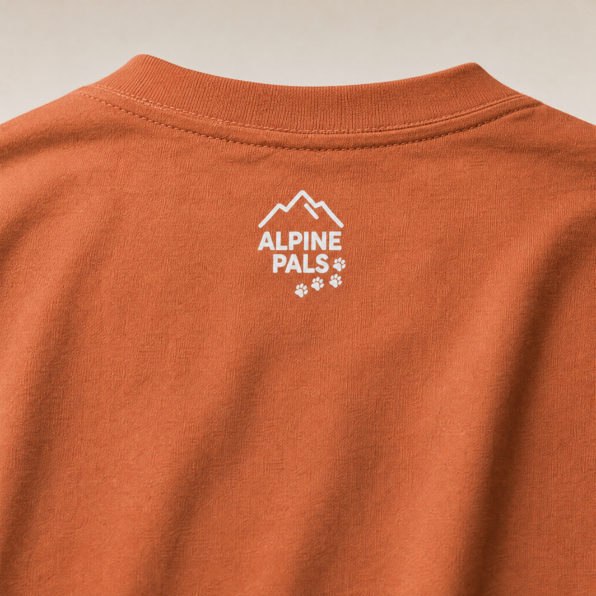 Close-up of a yam colored shirt with 'Alpine Pals' logo on a beige background