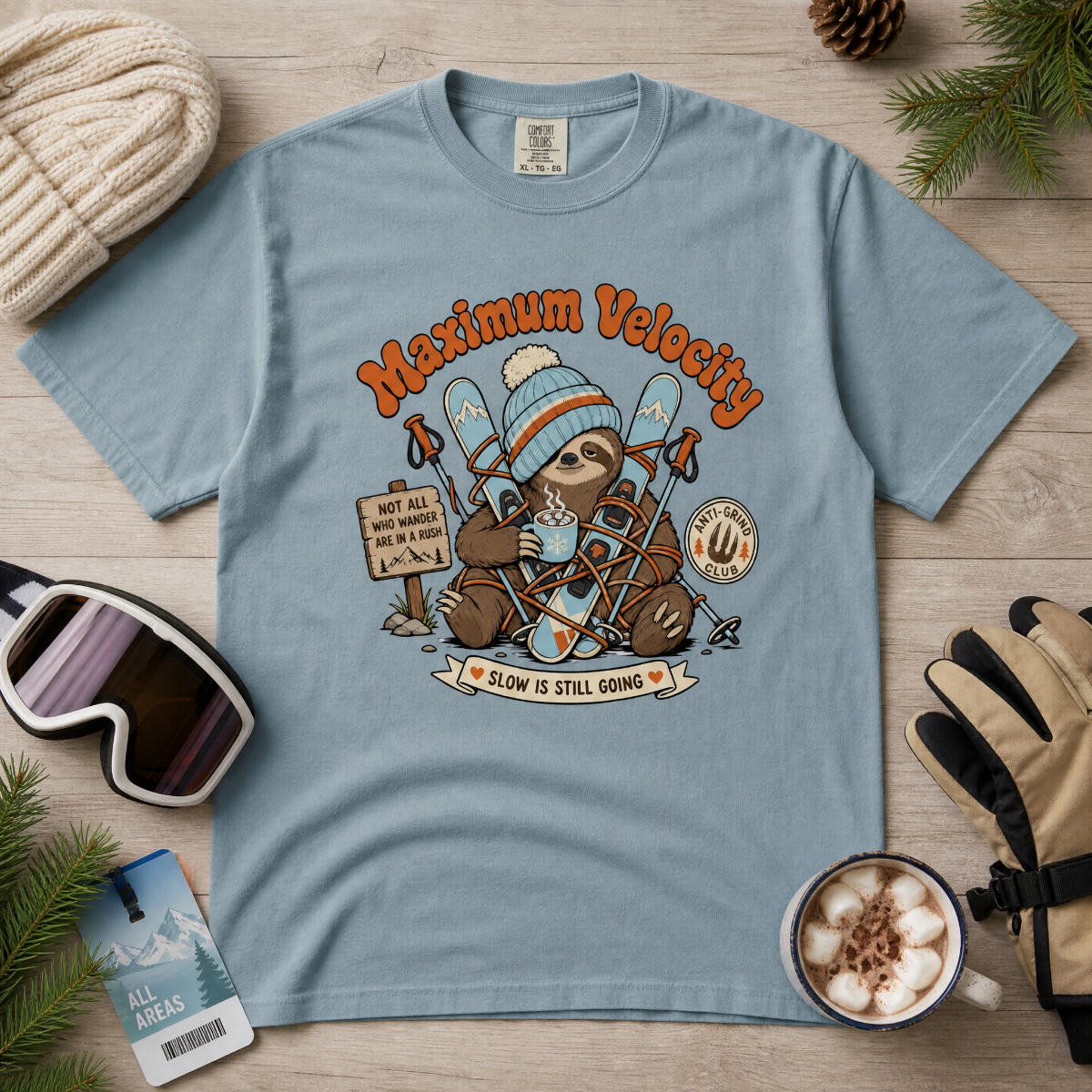 Ice blue colored t-shirt with a sloth graphic and 'Maximum Velocity' text on a wooden surface with winter accessories.
