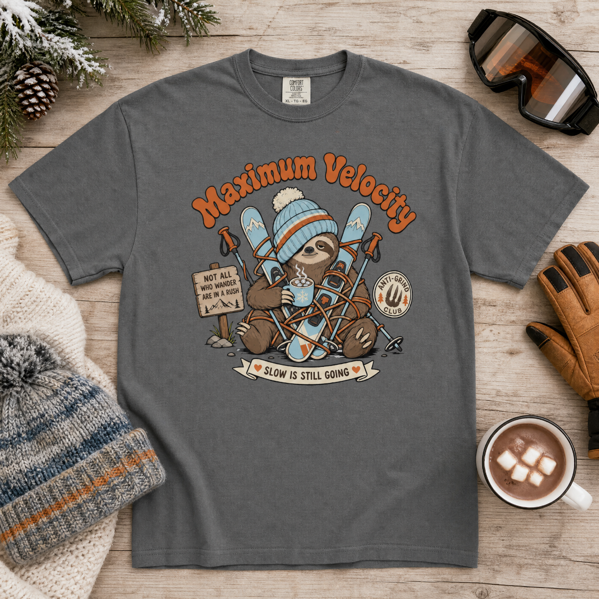 Pepper gray t-shirt with a sloth graphic and 'Maximum Velocity' text on a wooden surface with winter accessories.