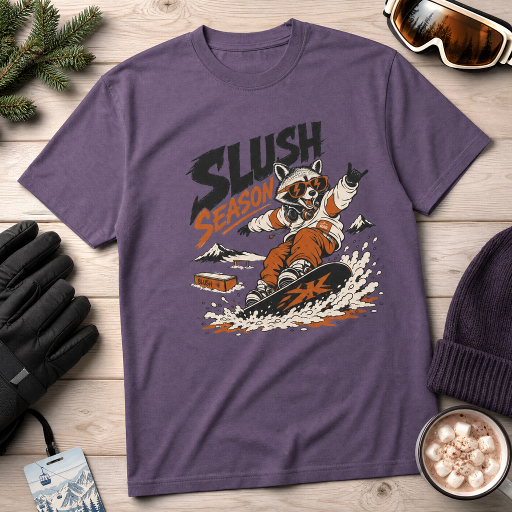 Purple t-shirt with a raccoon graphic and 'Slush Season' text on a wooden surface with winter accessories.