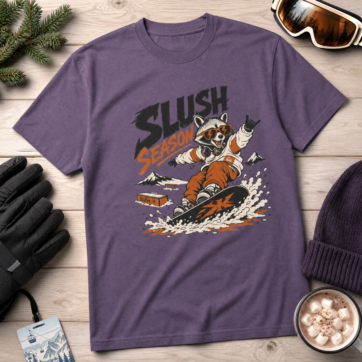 Purple t-shirt with a raccoon graphic and 'Slush Season' text on a wooden surface with winter accessories.