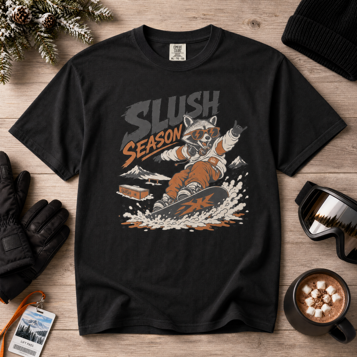 Black t-shirt with a raccoon graphic and 'Slush Season' text on a wooden surface with winter accessories.
