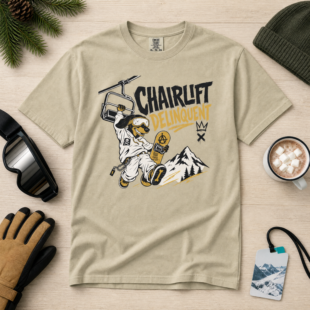 Khaki colored t-shirt with a snowboarding graphic and 'Chairlift Delinquent' text on a wooden surface with winter accessories.
