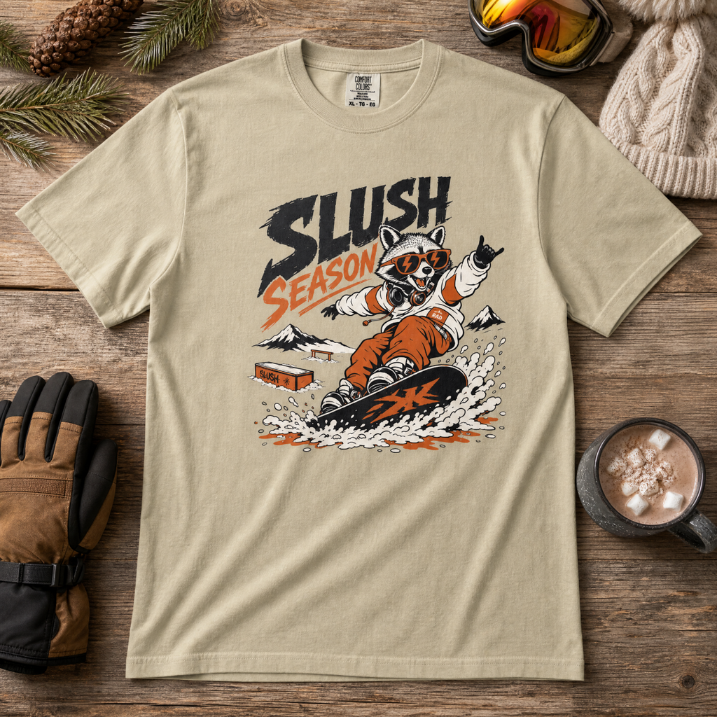 Khaki t-shirt with a graphic of a raccoon snowboarding and 'Slush Season' text, placed on a wooden surface with winter accessories.