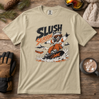 Khaki t-shirt with a graphic of a raccoon snowboarding and 'Slush Season' text, placed on a wooden surface with winter accessories.