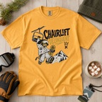 Mustard yellow t-shirt with a graphic design of a snowboarder and 'Chairlift Delinquent' text, surrounded by winter accessories on a wooden surface.