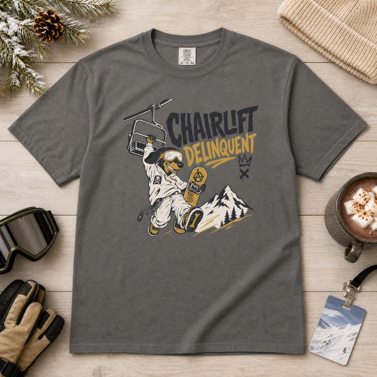 Pepper gray t-shirt with a graphic design of a snowboarder and 'Chairlift Delinquent' text on a wooden surface with winter accessories.