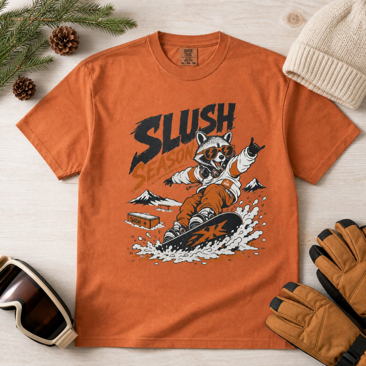 Yam colored t-shirt with a graphic of a raccoon snowboarding, surrounded by winter accessories on a light wooden surface.