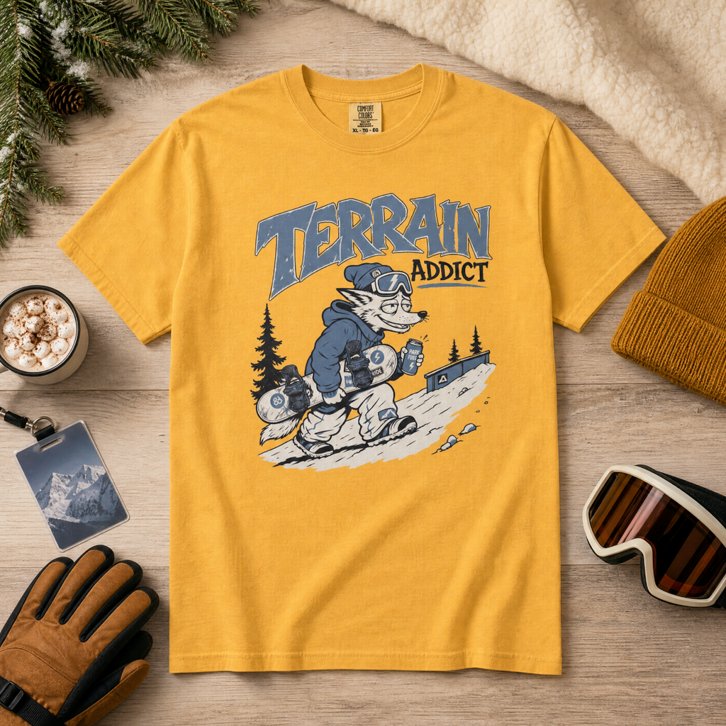 Mustard yellow t-shirt with 'Terrain Addict' graphic on a wooden surface with winter accessories.
