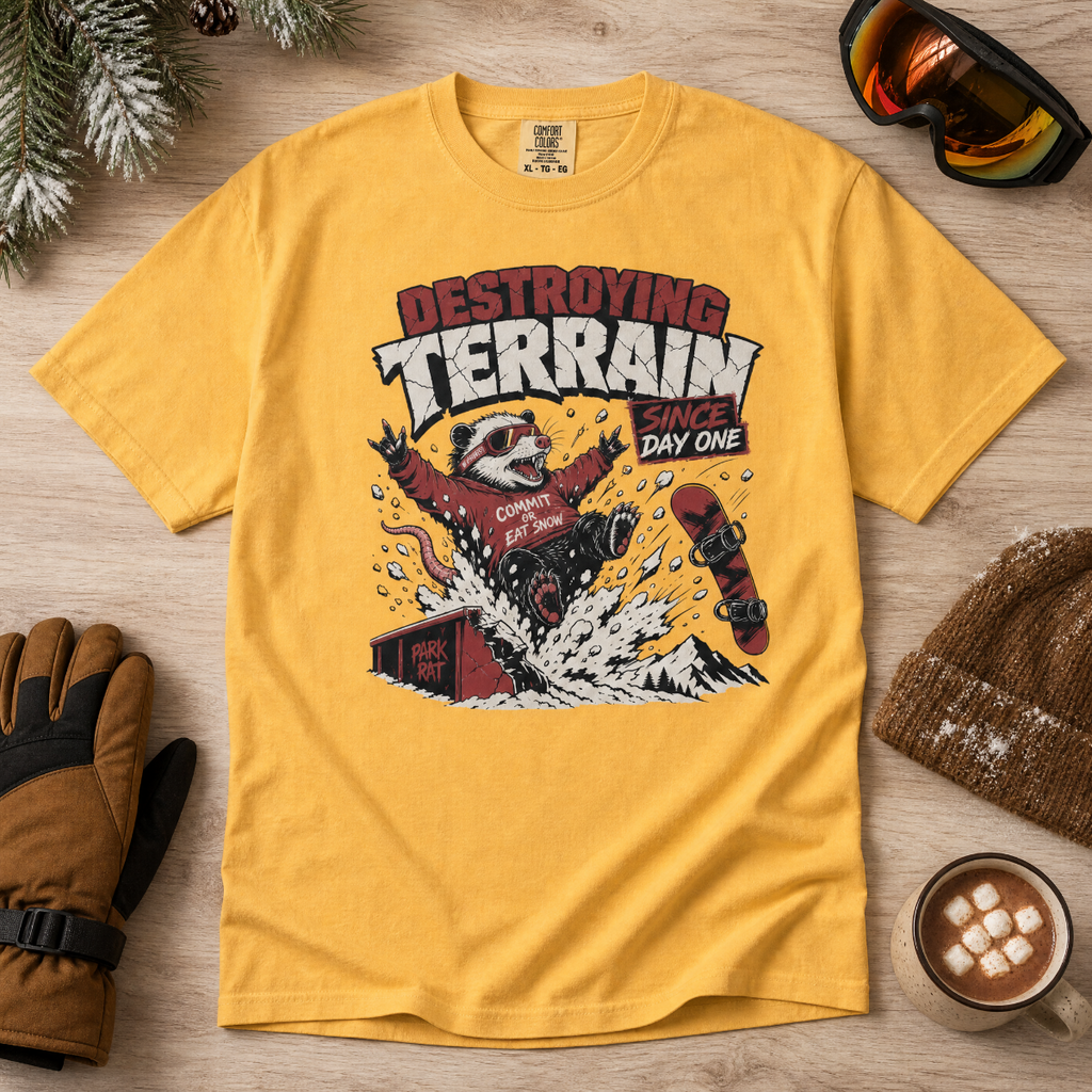 Mustard yellow t-shirt with a graphic design of a snowboarder and text 'Destroying Terrain Since Day One' on a wooden surface with winter accessories.
