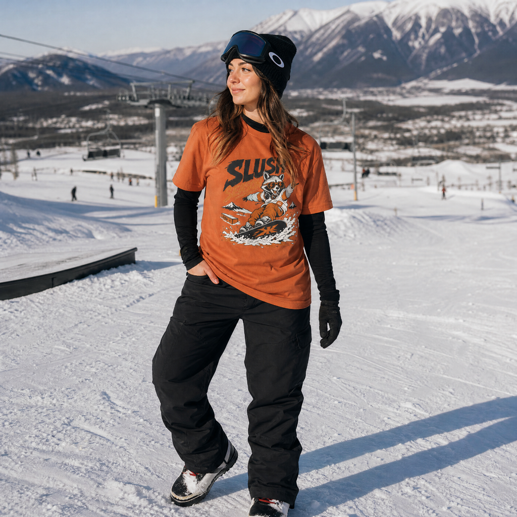 Person wearing an orange 'Slush Season' t-shirt and black pants on a snowy slope with mountains in the background
