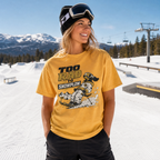 Person wearing a yellow t-shirt with a snowboarding graphic and text, standing in a snowy landscape with mountains and ski lifts.