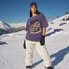 Person wearing a purple t-shirt with a graphic design, white pants, and black gloves standing on a snowy slope with mountains in the background.
