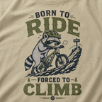 Khaki colored t-shirt with a graphic of a raccoon riding a bike and text 'Born to Ride, Forced to Climb'.