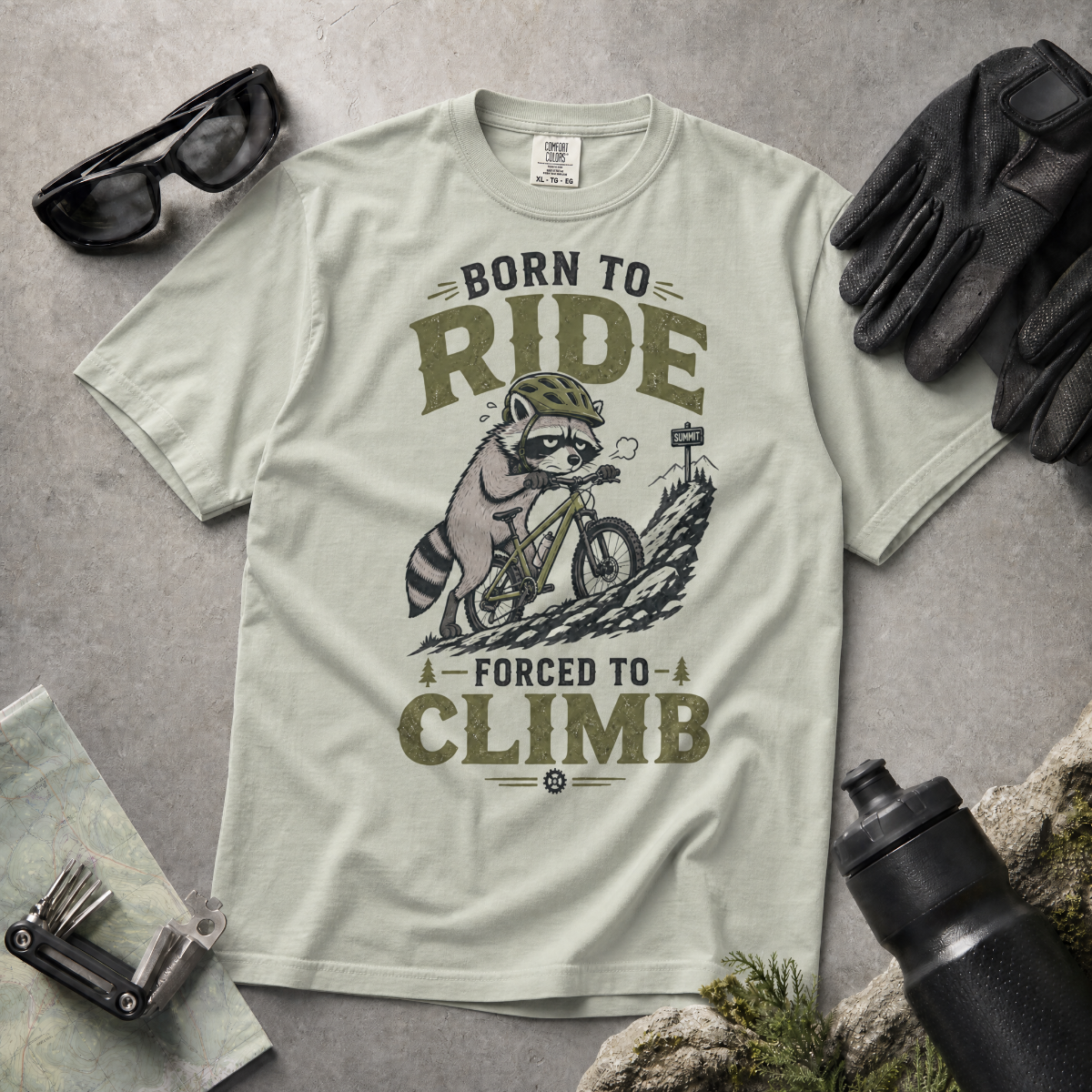 Bay green t-shirt with a graphic of a raccoon riding a bike and text 'Born to Ride, Forced to Climb'.