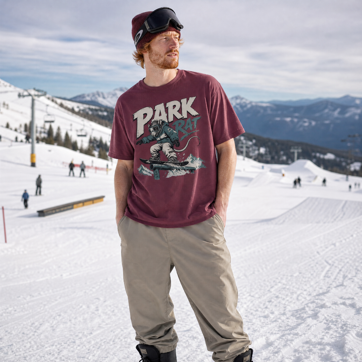Man wearing a Brick red t-shirt with 'Park Rat' graphic on a snowy slope with mountains in the background.