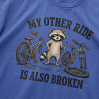 Mystic blue t-shirt with a raccoon and bicycle graphic and text 'My other ride is also broken' on a blue background.