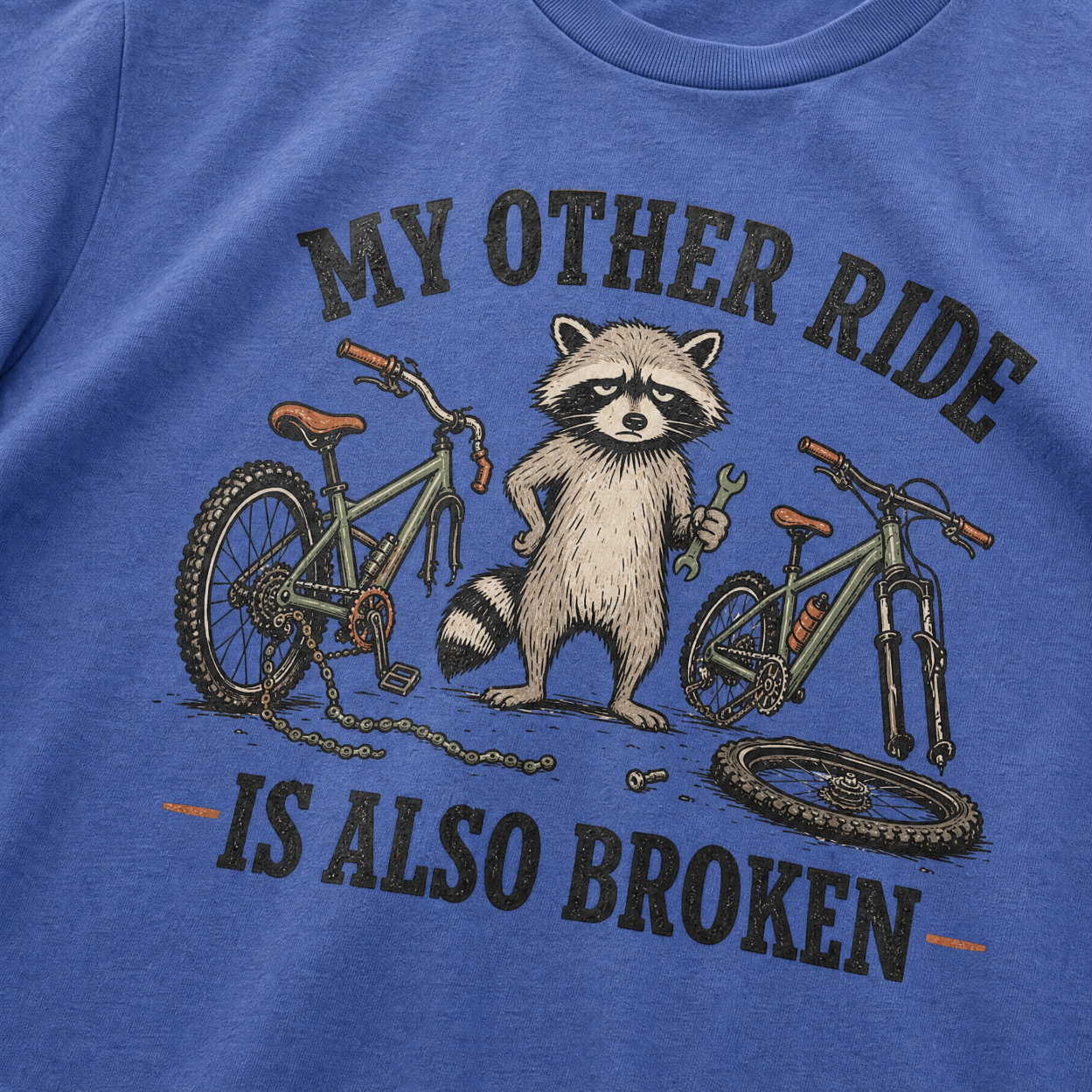 Mystic blue t-shirt with a raccoon and bicycle graphic and text 'My other ride is also broken' on a blue background.