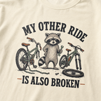 T-shirt with a raccoon and bicycles graphic and text 'My other ride is also broken' on a beige background.