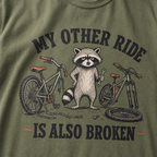 Moss green t-shirt with a raccoon graphic and text 'My other ride is also broken' on a green background.