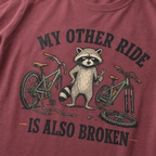 Brick red t-shirt with a raccoon and bicycle graphic and text 'My other ride is also broken' on a plain background.