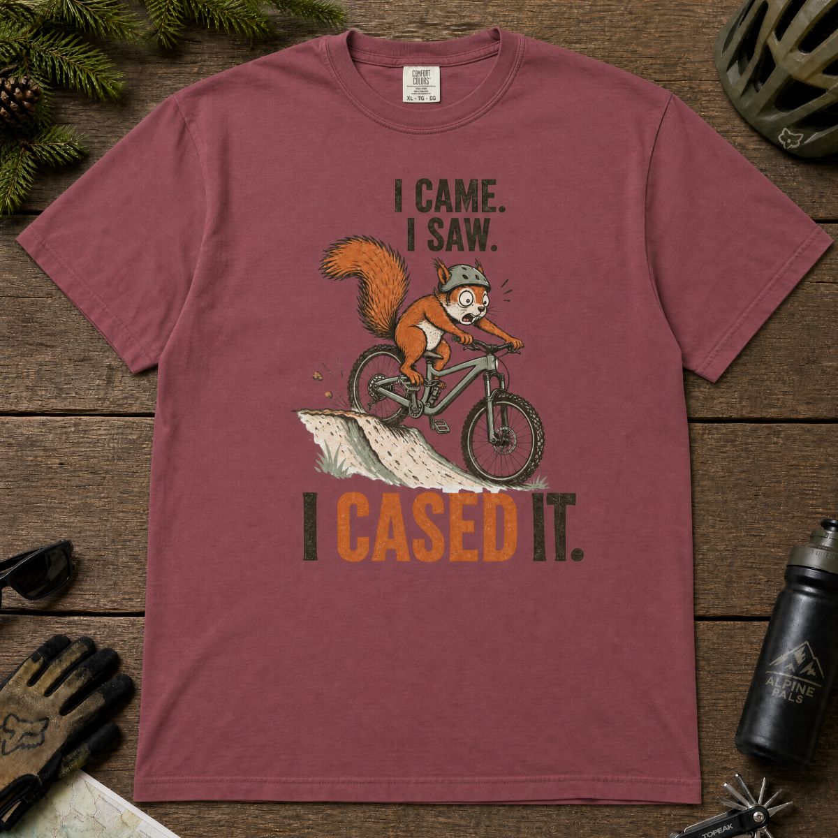 Brick red t-shirt with a graphic of a squirrel on a bike and text 'I came. I saw. I cased it.' on a wooden surface.