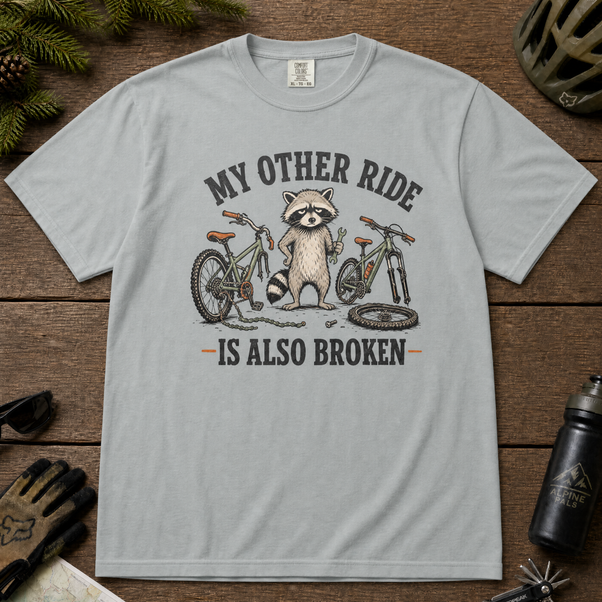 Granite gray t-shirt with a raccoon graphic and text on a wooden surface with outdoor gear.