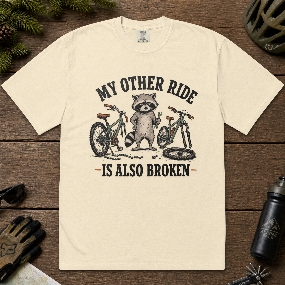 Ivory white t-shirt with raccoon graphic and text on a wooden surface with cycling accessories.